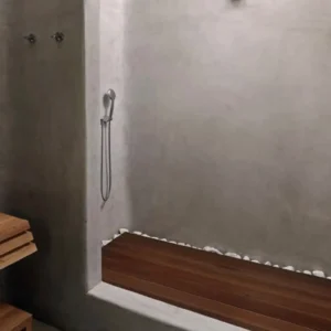 Develine Microcement gray shower enclosure with a teak wood bench, white river pebble floor border, and teak slatted vanity with white vessel sink