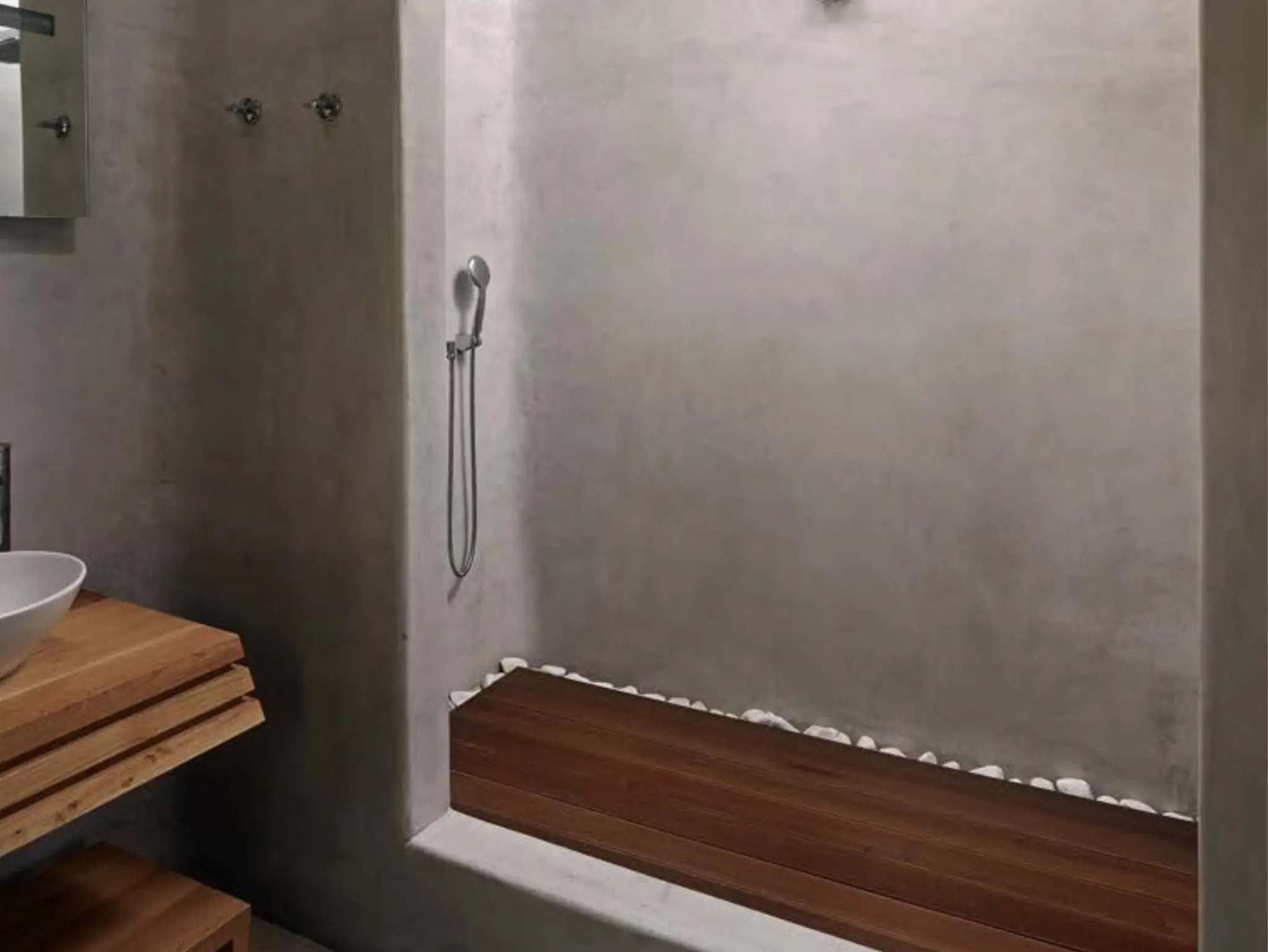Develine Microcement gray shower enclosure with a teak wood bench, white river pebble floor border, and teak slatted vanity with white vessel sink